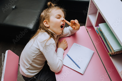 Tablou pe pânză Funny, lazy red-haired girl, child, schoolgirl sits at a wooden pink desk at school and picks her nose
