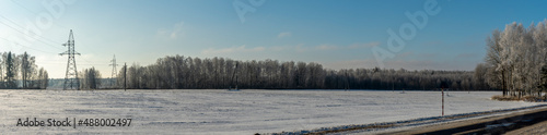 In background high-voltage electricity power pylon on snowy field with trees. High voltage poles on blue sky background.