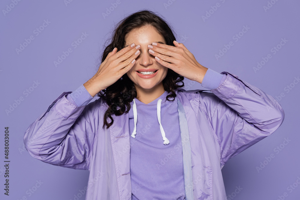 smiling young woman in raincoat covering eyes isolated on purple. smiling young woman in raincoat covering eyes isolated on purple.