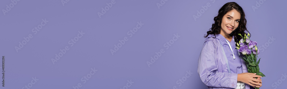 happy young woman in raincoat holding bouquet of flowers isolated on purple, banner. happy young woman in raincoat holding bouquet of flowers isolated on purple, banner.