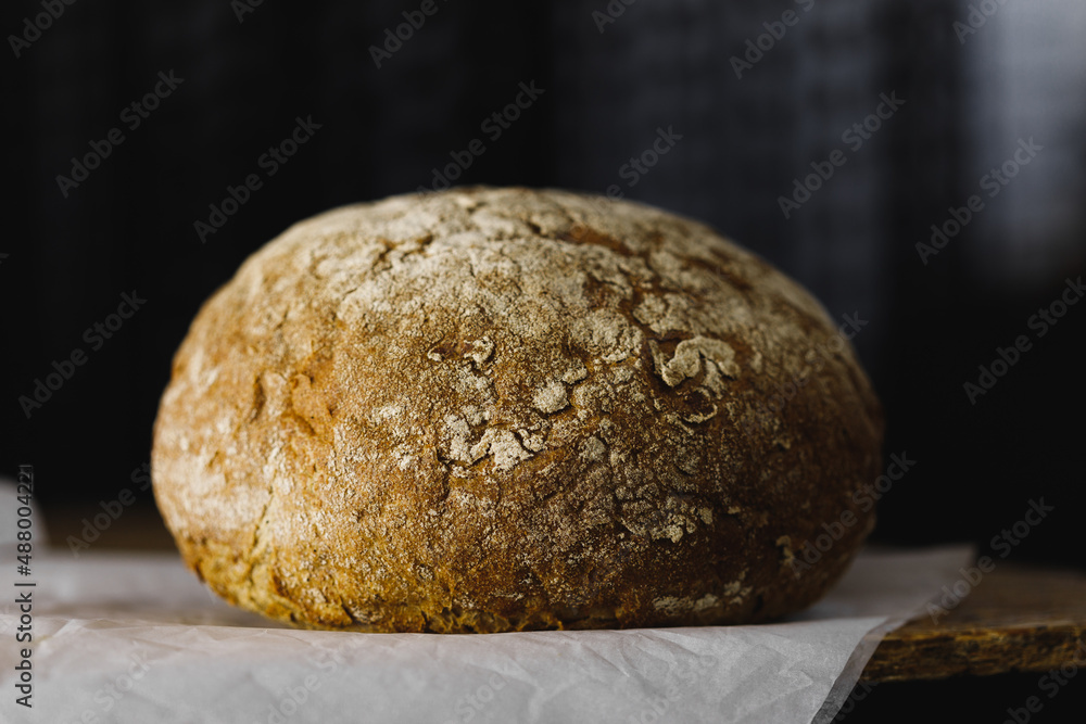 round bread with a bright crispy crust lies on parchment on a wooden table, dark background, side view