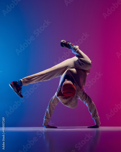 Studio shot of young flexible sportive man dancing breakdance in white outfit on gradient pink blue background. Concept of action, art, beauty, sport, youth