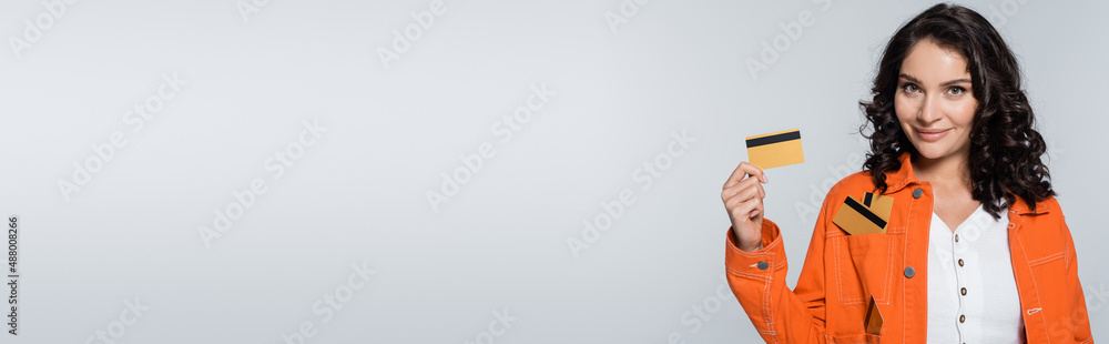 pleased young woman with credit cards in pocket of orange jacket isolated on grey, banner. pleased young woman with credit cards in pocket of orange jacket isolated on grey, banner.