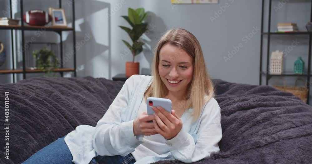 Close up portrait of pretty caucasian woman reclining on sofa texting via phone and laughing with teeth. Female scrolling social media on gadget. People after work resting on sofa with smartphones. Close up portrait of pretty caucasian woman reclining on sofa texting via phone and laughing with teeth. Female scrolling social media on gadget. People after work resting on sofa with smartphones.