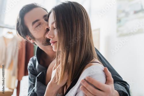 young man with closed eyes kissing cheek of pretty girlfriend in silk robe.