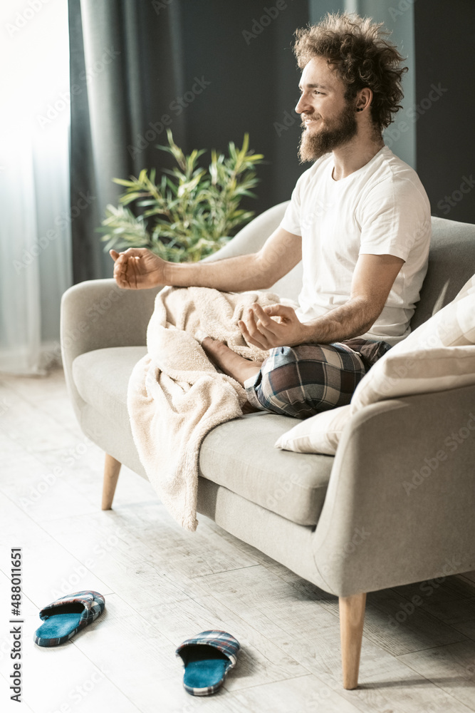 Handsome man sitting on the couch or sofa at home, enjoying meditation in his pajamas in living room during lockdown or being sick or weekend relax Handsome man sitting on the couch or sofa at home, enjoying meditation in his pajamas in living room during lockdown or being sick or weekend relax