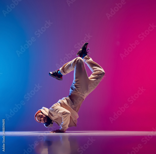 Free fall. Studio shot of young flexible sportive man dancing breakdance in white outfit on gradient pink blue background. Concept of action, art, beauty, sport, youth