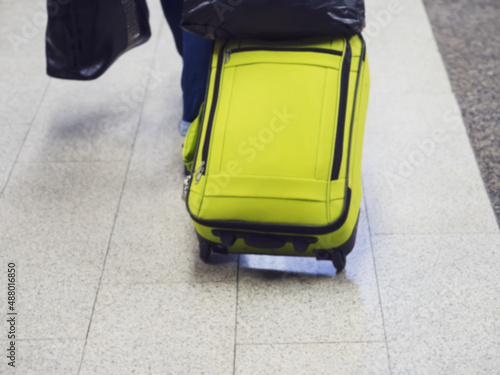 A passenger with a yellow suitcase on wheels walks on marble floor tiles. Defocused image.