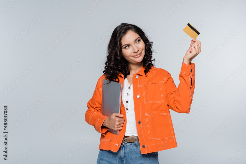 pleased woman in orange jacket holding laptop and looking at credit card isolated on grey. pleased woman in orange jacket holding laptop and looking at credit card isolated on grey.