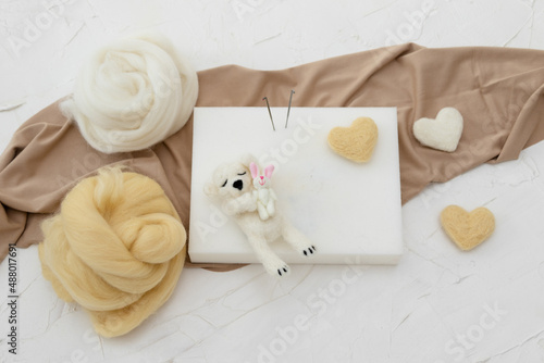 A small sleeping woolen white teddy bear made by dry felting. Felt toy for a newborn photo session. handmade product. Workplace for dry felting with a foam sponge, needles, fabric and skeins of wool 