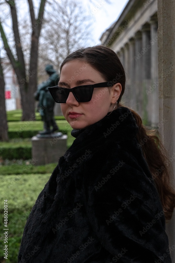 Portrait of a young woman in all-black outfit and black sunglasses on a grey cloudy day in park. Black clothes, typical Berlin. Green cut bushes, statue, columns