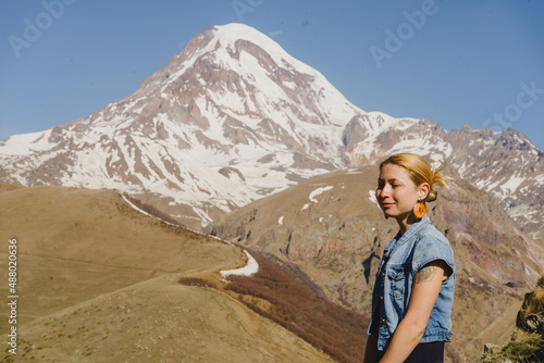 A young beautiful girl in a blue long dress and a denim jacket is sitting against the backdrop of the high Kazbek mountains in the rays of the dawn sun. Georgia. Europe