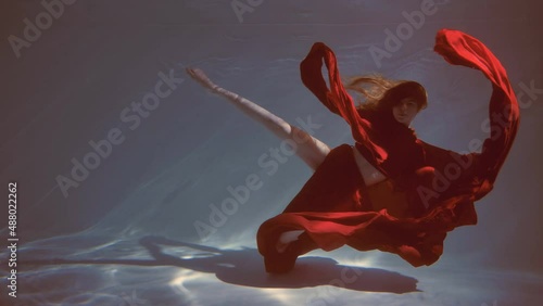 Attractive red-haired young woman swims beautifully underwater in a red dress. slow motion