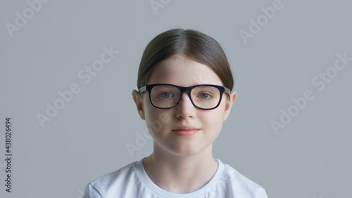 A blonde teenager girl in a white T-shirt puts on and takes off red glasses against the background of a white wall and smiles
