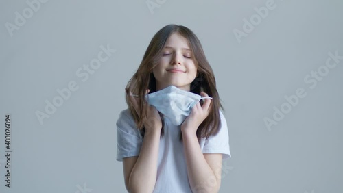 A blonde teenager girl in a white T-shirt takes off a blue mask against the background of a white wall and smile