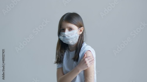 A blonde teenager girl in a white t-shirt and a blue mask touches the vaccine on her shoulder in front of the background of a white wall