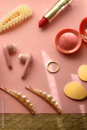 Pink bluetooth earphones, red hairband, pearl hair clips, gold earrings and ring, pink blush and lipstick on pastel pink background. Selective focus.