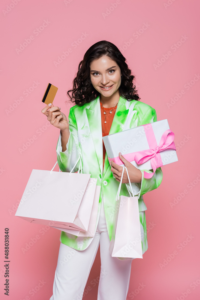 cheerful young woman in tie dye blazer holding credit card, shopping bags and wrapped present isolated on pink. cheerful young woman in tie dye blazer holding credit card, shopping bags and wrapped present isolated on pink.