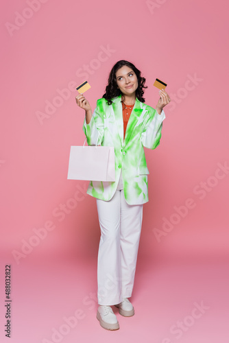 full length of smiling woman in tie dye blazer holding credit cards and shopping bag on pink.