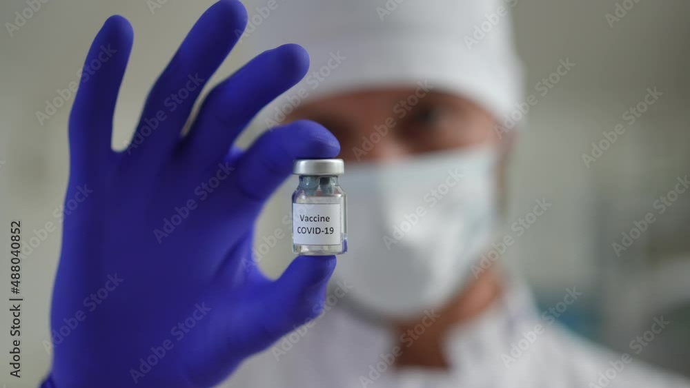 Rack focus from serious doctor scientist in coronavirus face mask to Covid-19 vaccine jab in hand. Confident Caucasian man posing in vaccination clinic with virus cure. Health care and medicine Rack focus from serious doctor scientist in coronavirus face mask to Covid-19 vaccine jab in hand. Confident Caucasian man posing in vaccination clinic with virus cure. Health care and medicine