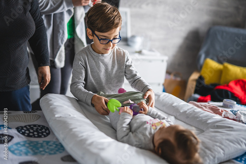 Caucasian boy little child taking care of his baby brother or sister changing clothes getting dressed while lying on the bed at home in bright day real people family and bonding concept copy space