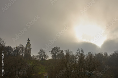 There is a tall bell tower on the hill against a cloudy sky . The sun peeks through the clouds.