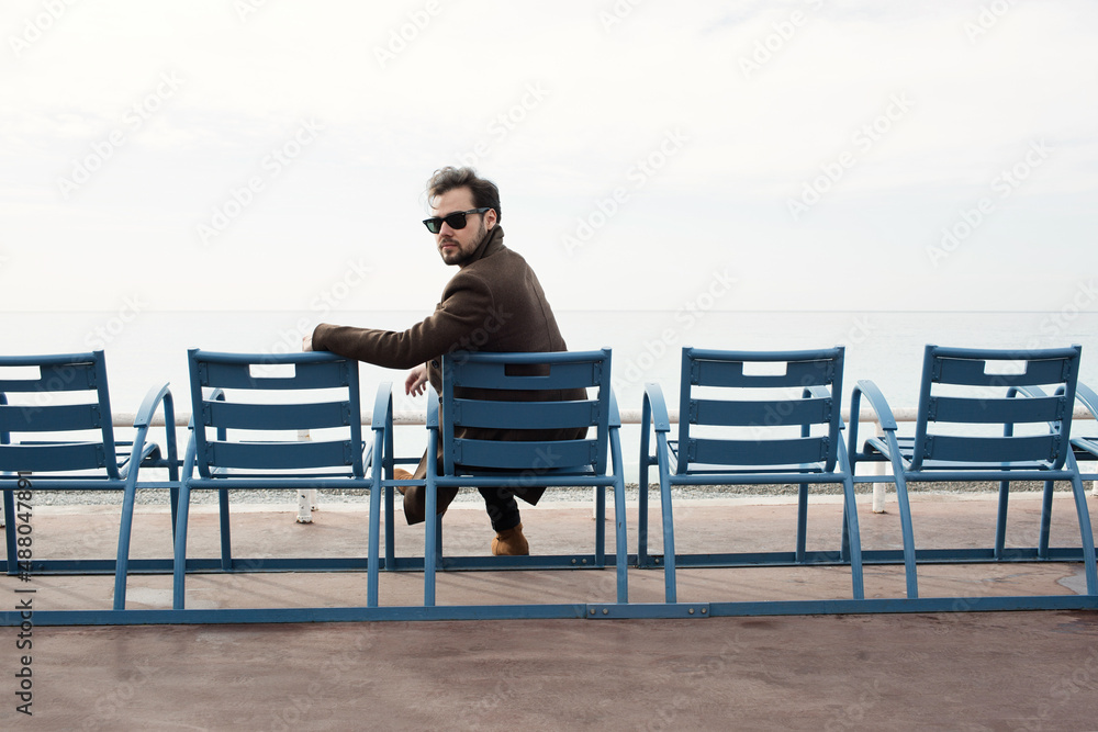 Bearded man in coat sitting on the bench against the sea coast. 