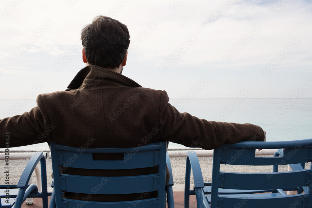 Fototapeta premium Back view of man in coat sitting on the bench against the sea coast. Cropped.