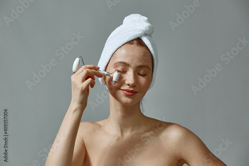 Young girl in towel massage face by jade roller massager to improve complexion, blood circulation, enjoying skin care