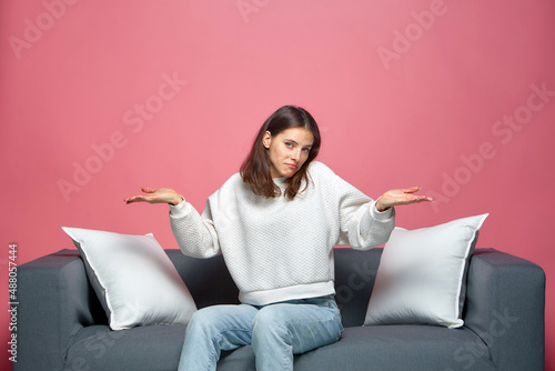Puzzled young woman feel perplexity shrugged shoulders, spreading hands, sitting on sofa. I dont know. Misunderstanding