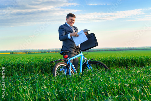 businessman dressed in a business suit, poses with bicycle in green grass field and reads documents or reports, beautiful nature in spring, business concept © soleg
