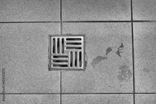 Metal drain hole in the tiled floor of a shower. Floor drain, running water in shower, tinted black and white image