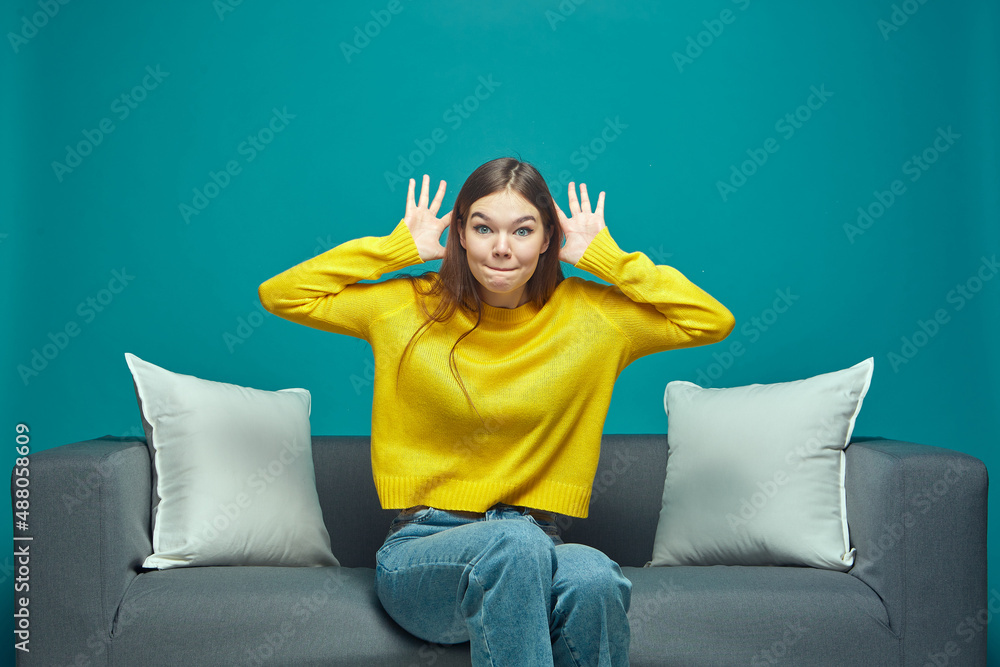 Funny young girl making faces, grimacing, fooling, showing big ears by hands, teasing, sitting on sofa Funny young girl making faces, grimacing, fooling, showing big ears by hands, teasing, sitting on sofa