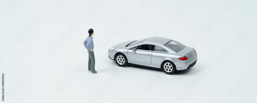 On a white surface is a car and a miniature figurine of a man looking at the car.