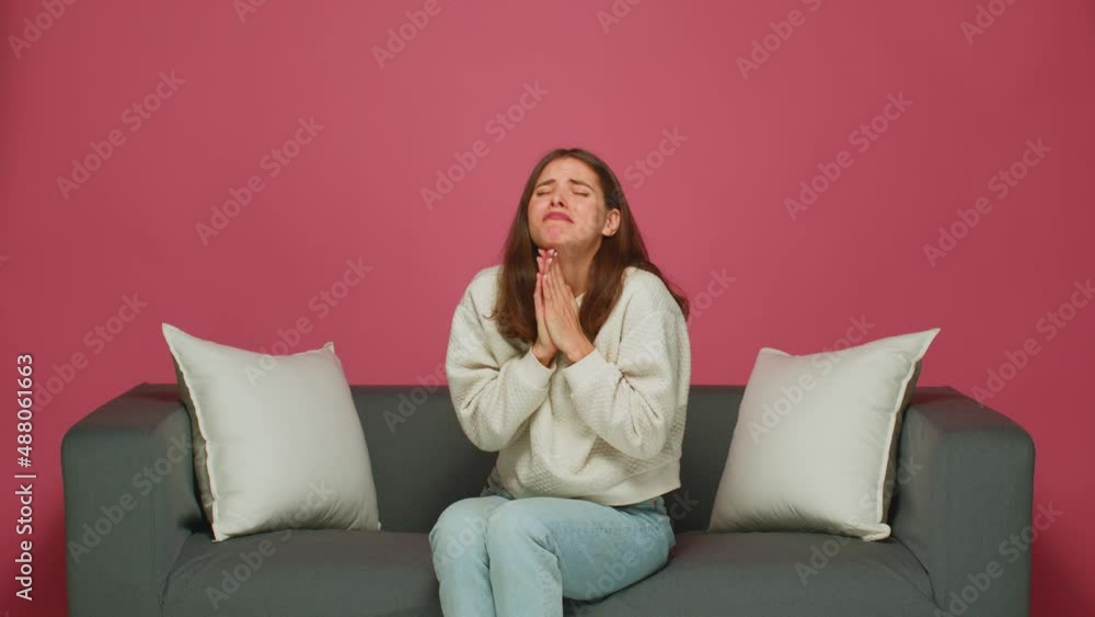 Young girl begging, pleading folded hands asking help or forgiveness, sitting on sofa on pink studio background Young girl begging, pleading folded hands asking help or forgiveness, sitting on sofa on pink studio background