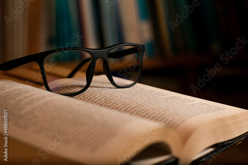 Eye glasses on top of an open book. Literature day and knowledge concept. Blurred bookshelf on the background. Selective focus.
