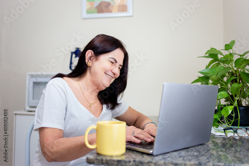 Mature woman using laptop at home to browse the internet or work, with a cup of coffee by her side.
