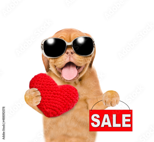 Happy mastiff puppy wearing sunglasses holds heart shaped balloon and sales symbol. isolated on white background