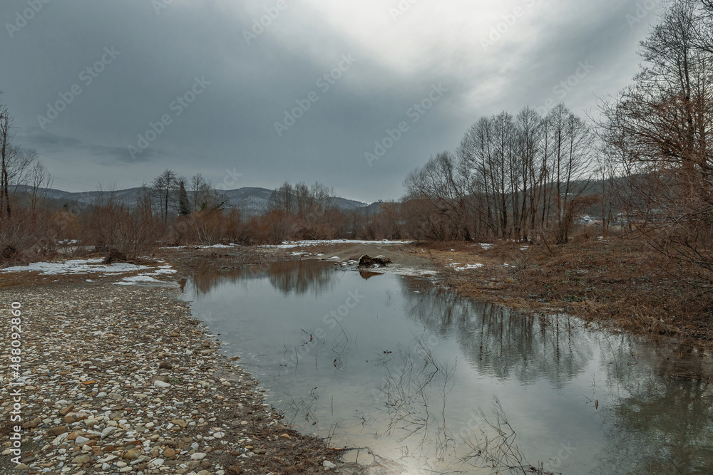 Reflection of the sky and clouds in a puddle. The gloomy winter sky is reflected in the water of melting snow. A rare forest with a mountain river in winter on a cloudy day. A cloudy day in the foothi