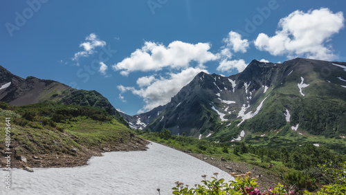 A picturesque mountain range against a background of blue sky and clouds. On the slopes there is green vegetation and melted snow. Kamchatka. Vachkazhets 