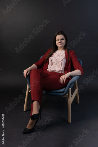 Cute plus-size model is sitting on a chair on a black background in an unbuttoned red jacket, a pink blouse, black trousers and high-heeled shoes. Business portrait. Business woman in red suit