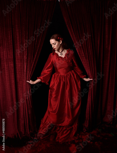   portrait of pretty female model with red hair wearing glamorous historical victorian red ballgown.  Posing with a moody dark background.