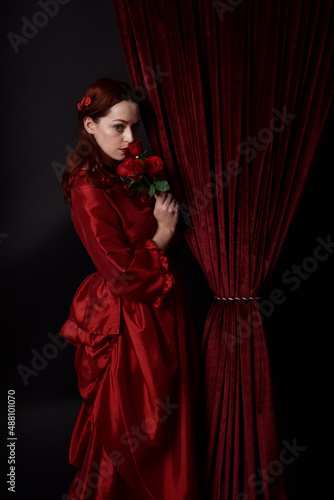  portrait of pretty female model with red hair wearing glamorous historical victorian red ballgown. Posing with a moody dark background.