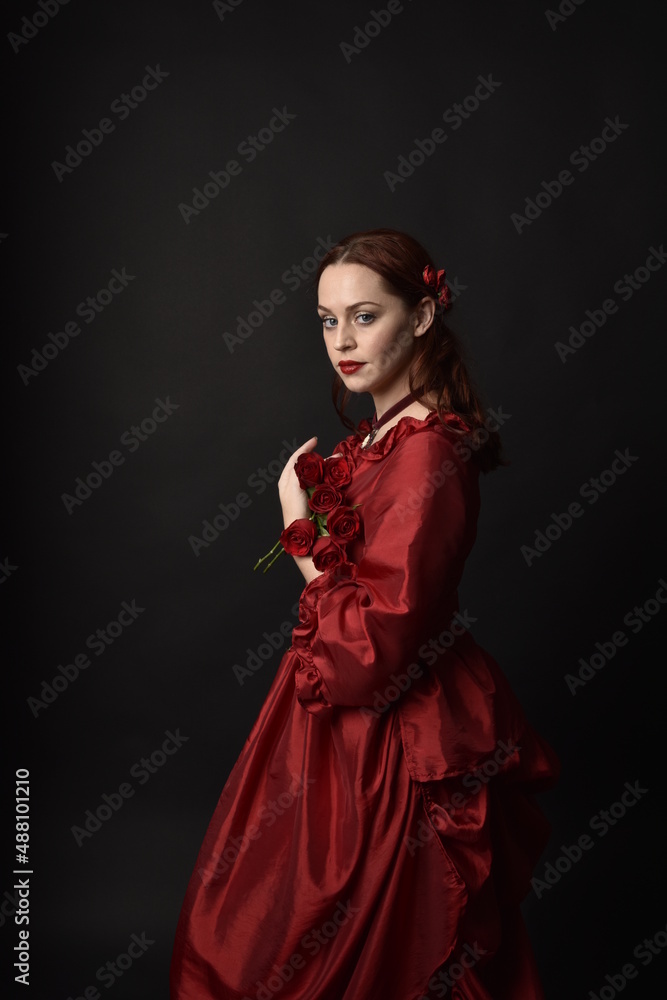 portrait of pretty female model with red hair wearing glamorous historical victorian red ballgown. Posing with a moody dark background. portrait of pretty female model with red hair wearing glamorous historical victorian red ballgown. Posing with a moody dark background.