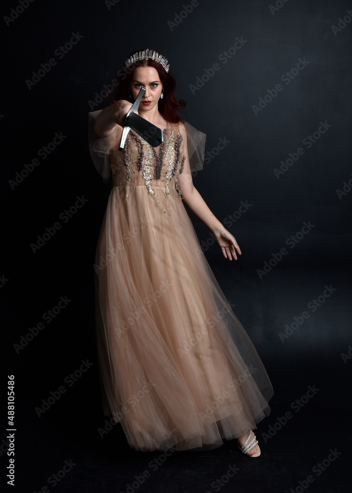  Full length portrait of pretty female model with red hair wearing glamorous fantasy tulle gown and crown.  Posing with a moody dark background.