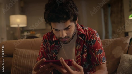 track in shot of a young Indian Asian Male guy sitting on a couch watching a live telecast match of football or cricket on a Mobile phone in an interior house setup. concept of entertaining sports.