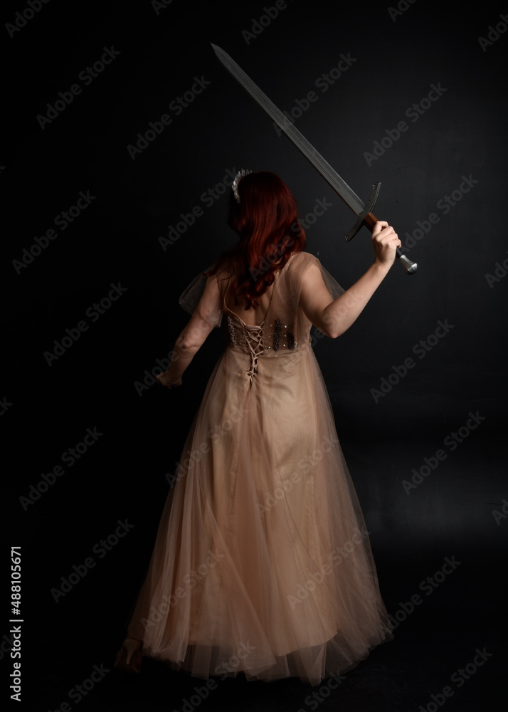 Full length portrait of pretty female model with red hair wearing glamorous fantasy tulle gown and crown. Posing with a moody dark background. Full length portrait of pretty female model with red hair wearing glamorous fantasy tulle gown and crown. Posing with a moody dark background.
