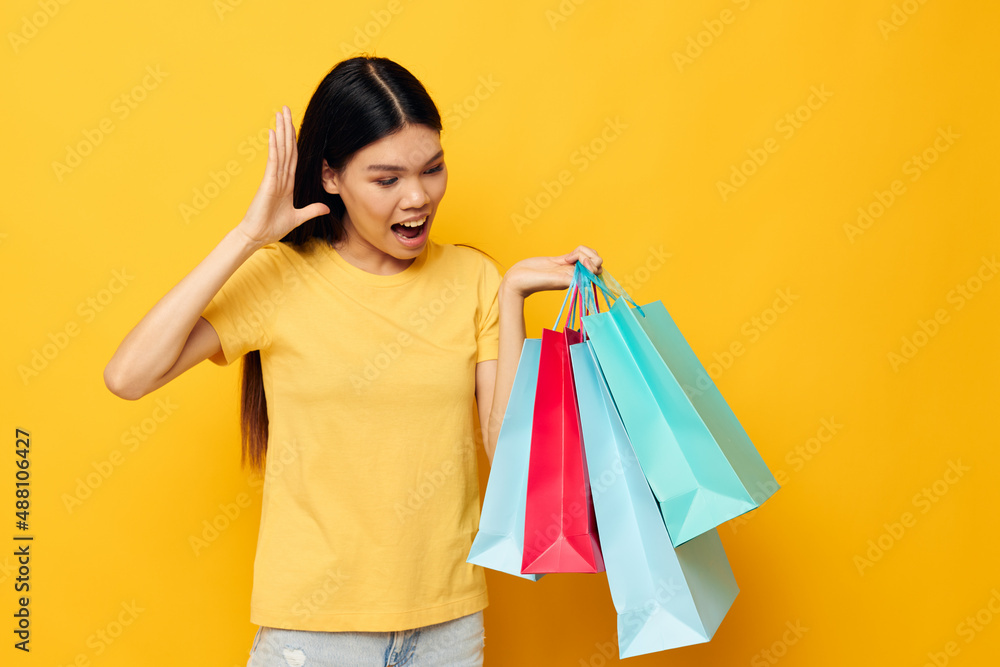 Charming young Asian woman in a yellow T-shirt with multicolored shopping bags yellow background unaltered Charming young Asian woman in a yellow T-shirt with multicolored shopping bags yellow background unaltered