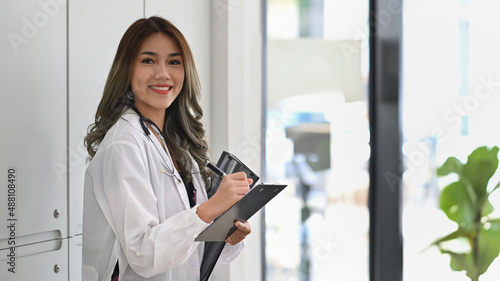 Beautiful female doctor in white coat with stethoscope holding her patient chart and smiling to camera.