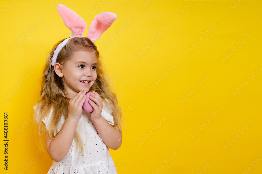 beautiful smiling blonde girl with bunny ears holding an easter egg in her hands on a yellow background, kid celebrate easter. beautiful smiling blonde girl with bunny ears holding an easter egg in her hands on a yellow background, kid celebrate easter.